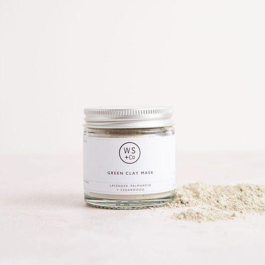 A clear jar containing green clay powder, with a label that reads 'Green Clay Mask'. The jar is placed on a light background, and there is some clay powder scattered in front of it.