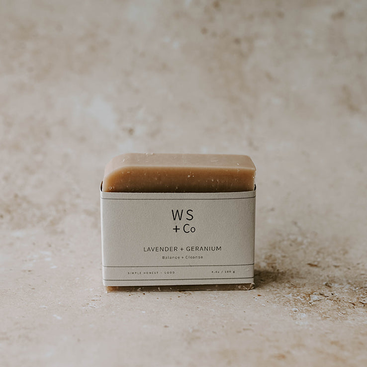 A bar of soap with a beige tone, packaging labeled 'WS + Co LAVENDER + GERANIUM', placed on a textured background.