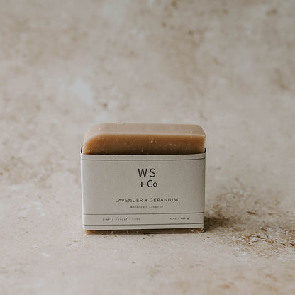 A bar of soap with a beige tone, packaging labeled 'WS + Co LAVENDER + GERANIUM', placed on a textured background.