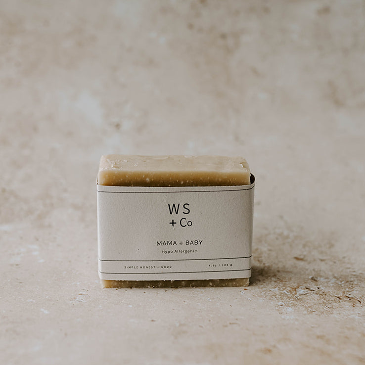A bar of handmade soap with a label that reads 'Mama + Baby Soap' and the brand 'Wild Sage + Co'. The soap is on a textured background.