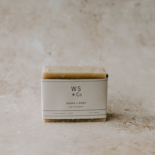 A bar of handmade soap with a label that reads 'Mama + Baby Soap' and the brand 'Wild Sage + Co'. The soap is on a textured background.