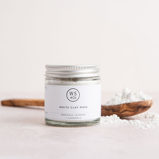 A clear glass jar containing white clay face mask, with a wooden spoon in the foreground.