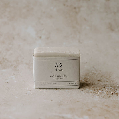 A bar of soap with 'WS + Co PURE OLIVE OIL' printed on it, placed on a textured background.