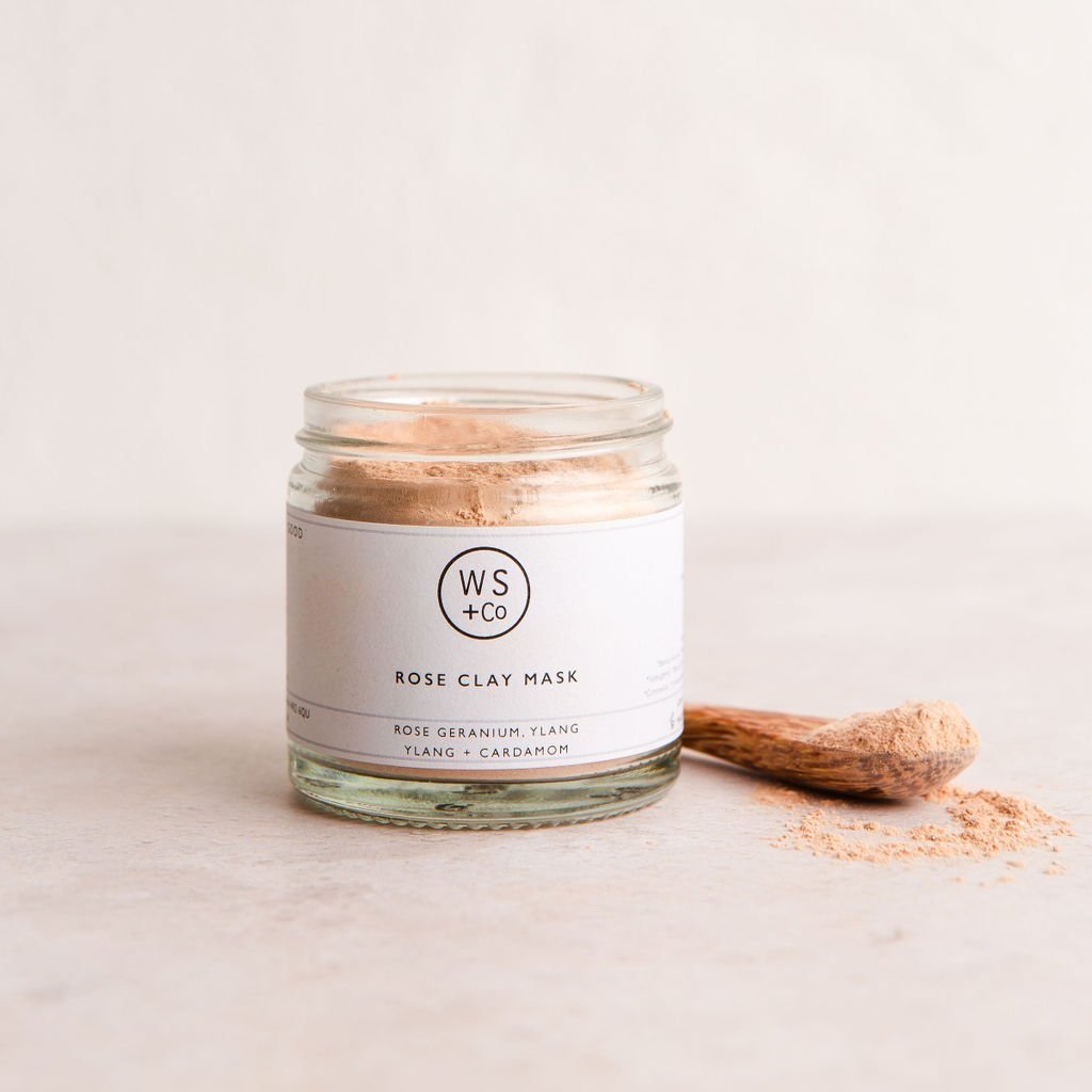 A glass jar containing rose clay powder, labeled 'ROSE CLAY FACE MASK' with a spoonful of the product in front, on a light background.