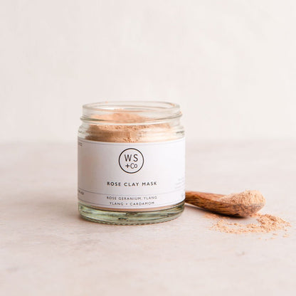 A glass jar containing rose clay powder, labeled 'ROSE CLAY FACE MASK' with a spoonful of the product in front, on a light background.