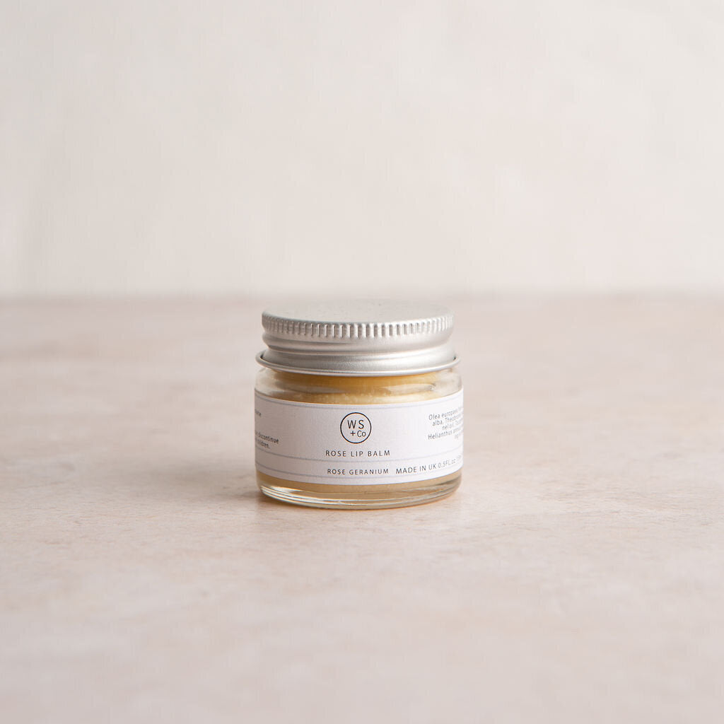 A small glass jar with a metal lid containing rose tinted lip balm, labeled with the brand name and product details.