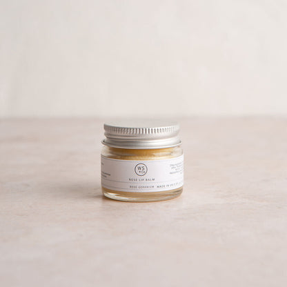 A small glass jar with a metal lid containing rose tinted lip balm, labeled with the brand name and product details.