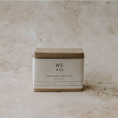A bar of soap with a label that reads 'WS + CO Rosemary Tea Tree' placed on a textured surface with a neutral background.