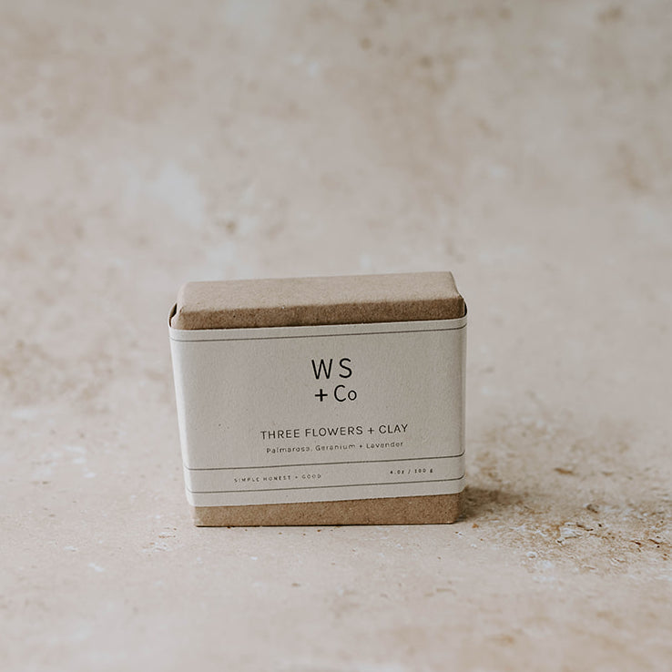 A bar of handmade soap inside a paper packaging labeled 'Three Flowers + Clay' by Wild Sage + Co. The packaging is brown with a minimalist design.