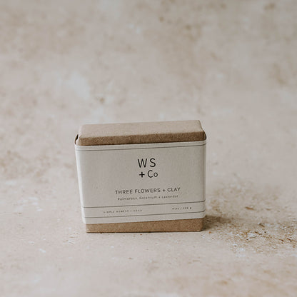 A bar of handmade soap inside a paper packaging labeled 'Three Flowers + Clay' by Wild Sage + Co. The packaging is brown with a minimalist design.